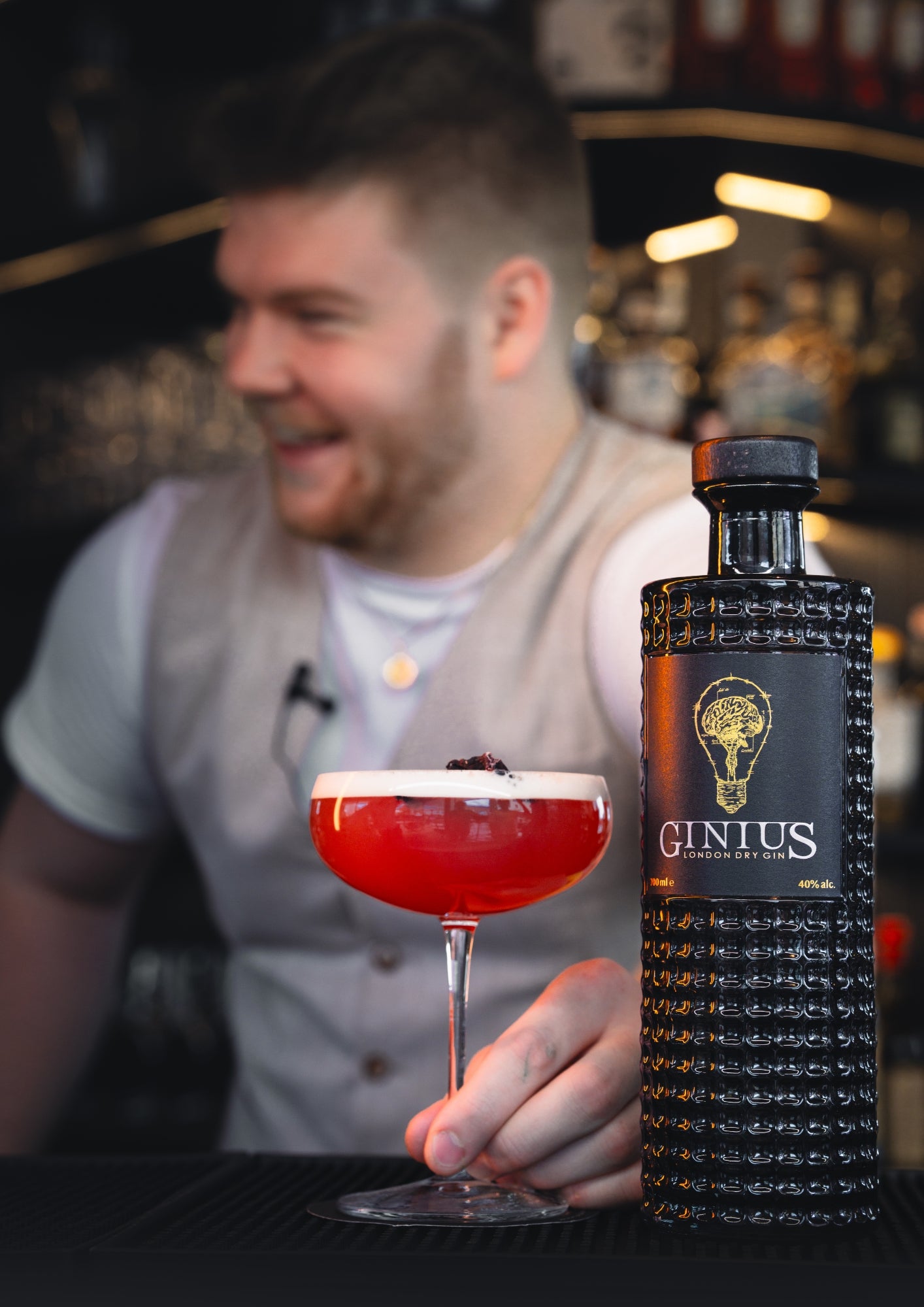 Person holding a red cocktail with a bottle of GINIUS in a bar setting. Martini glass